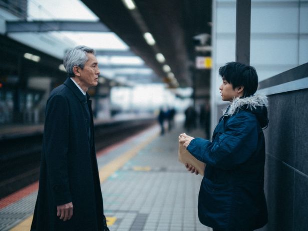 駅のホームで、謎の男性(松重)からなにかを受け取る佐野